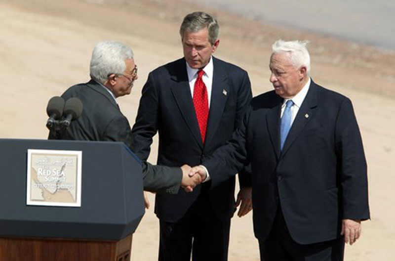Palestinian Authority leader Mahmoud Abbas, US President George W. Bush, and Israeli Prime Minister Ariel Sharon greet each other on stage behind a podium.