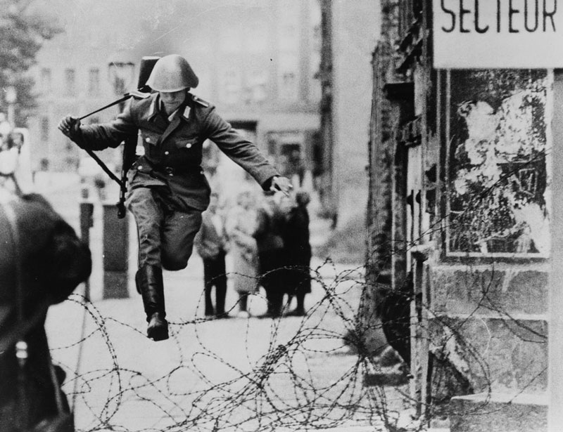 A black and white photo of an East German soldier defecting by jumping over barbed wire in the early days of the Berlin Wall. 