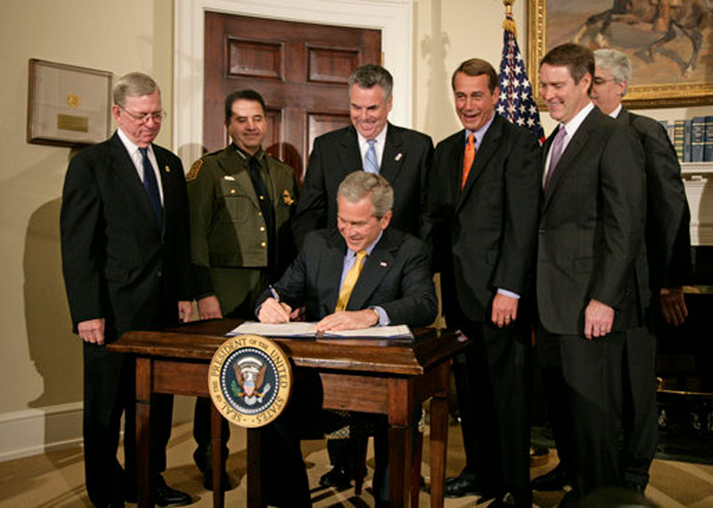 Former United States President George W. Bush sits at a desk and signs the Secure Fence Act of 2006 with members of Congress standing behind him.